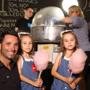 Father and two girls in denim overalls holding pink fairy floss at a Sarina Sugar Shed family hands-on sugar experience